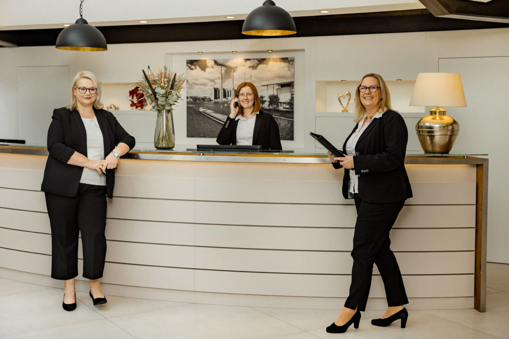 Drei Frauen in schwarzer Businesskleidung stehen an einer modernen Rezeption, eine telefoniert, eine hält einen Ordner, eine lehnt an der Theke.