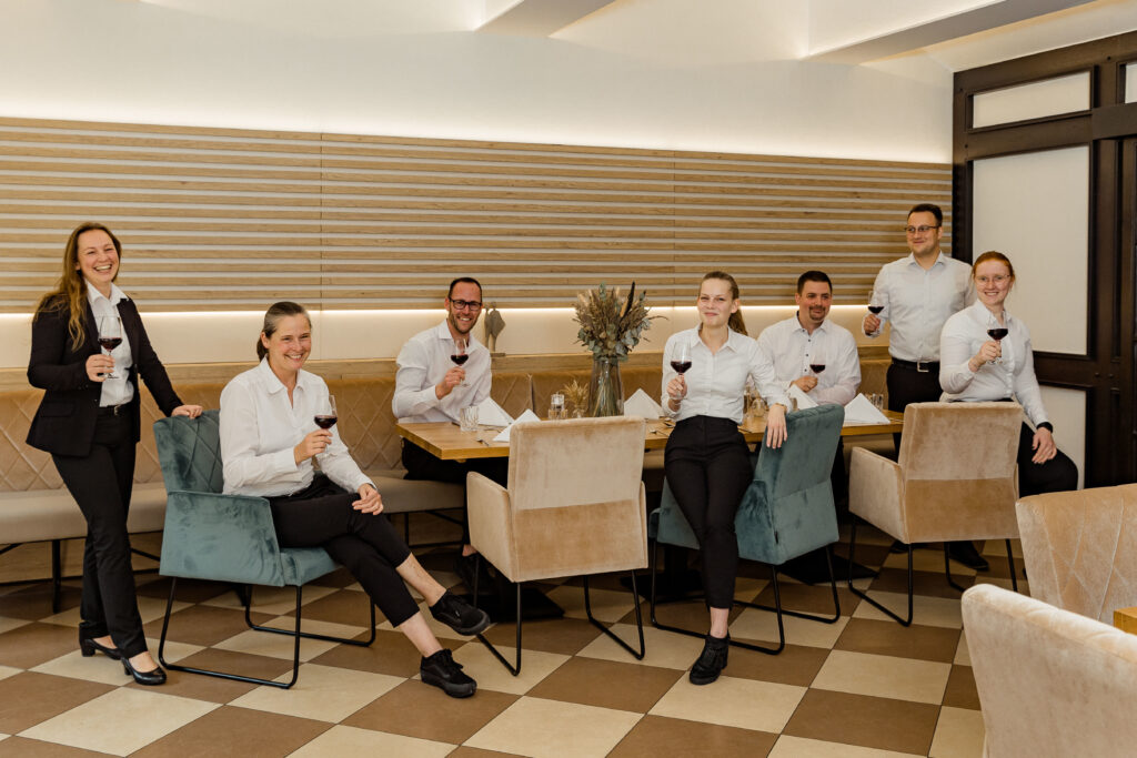 Sieben Personen in formeller Kleidung sitzen und stehen um einen Tisch in einem Restaurant und halten Rotweingläser zum Anstoßen.
