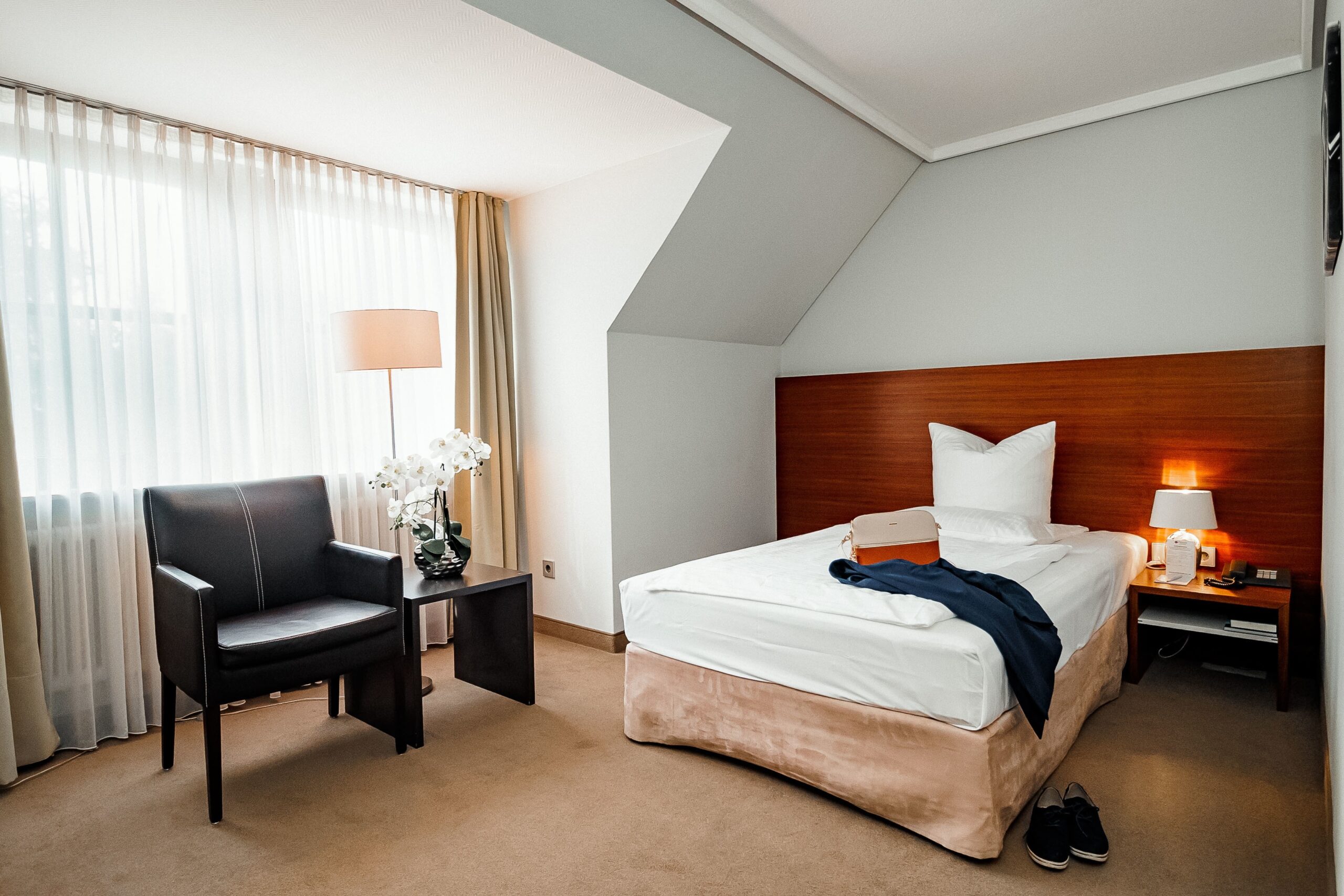 Standard-Einzelzimmer mit Einzelbett, Nachttisch mit Lampe, schwarzem Sessel, Beistelltisch und Fenster mit Vorhängen