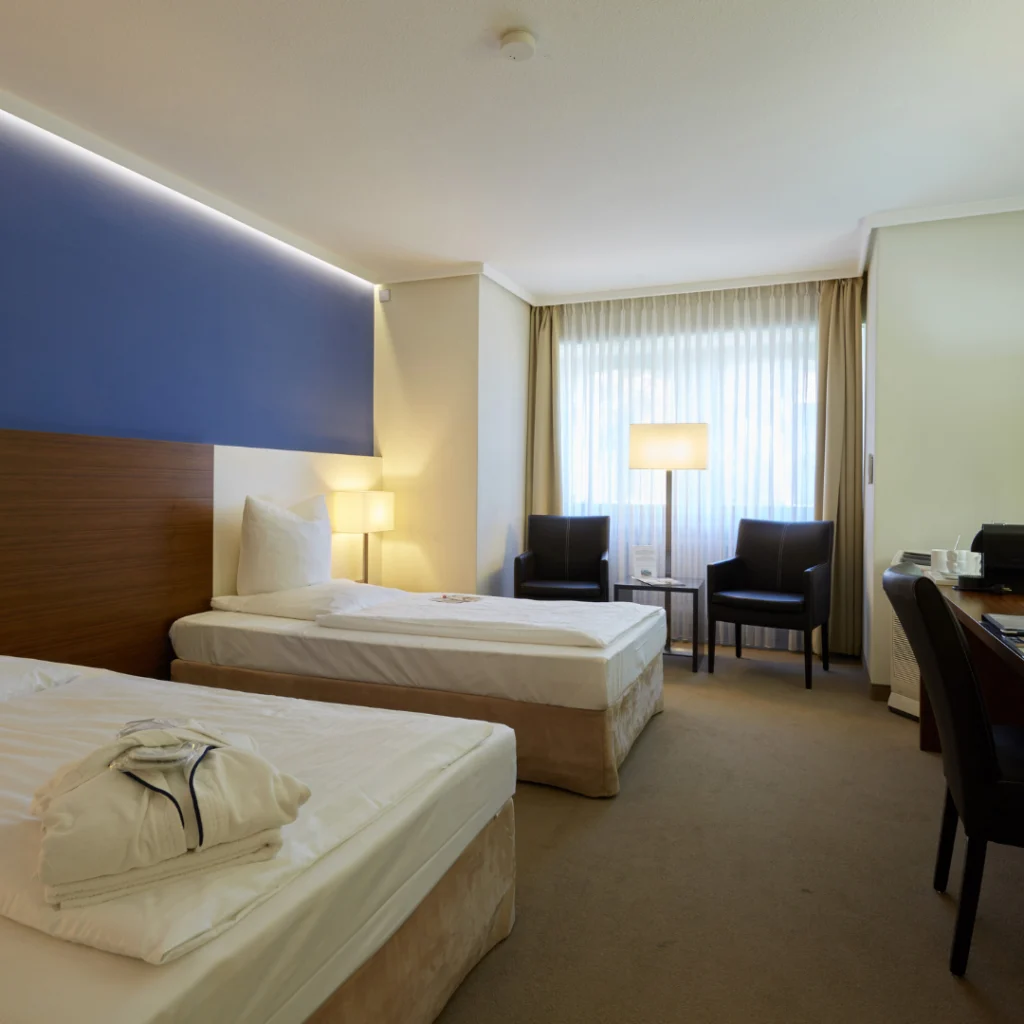 Standard-Doppelzimmer im Privathotel Alte Mühle mit zwei Einzelbetten, Nachttischlampen, zwei schwarzen Sesseln vor Fenster mit Vorhängen und Schreibtisch mit Stuhl.