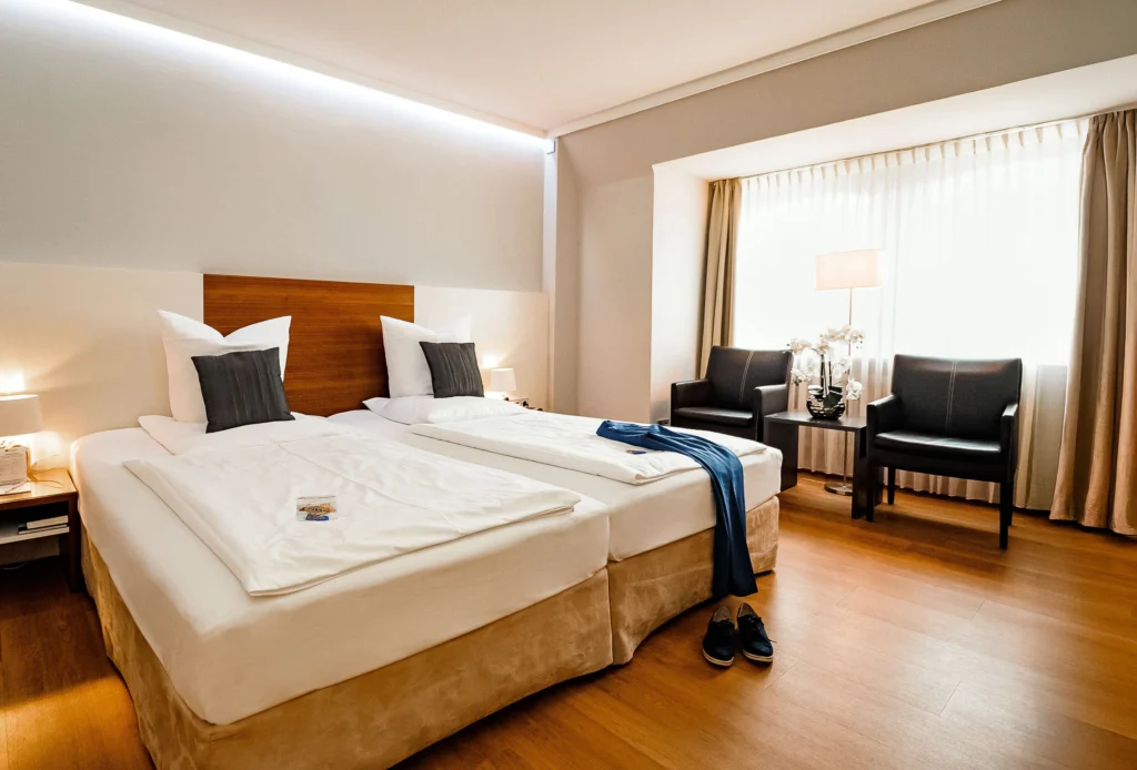 Zimmer im Privathotel ALTE MÜHLE mit zwei Einzelbetten, Holzboden, zwei schwarzen Sesseln am Fenster und Nachttischen mit Lampen.