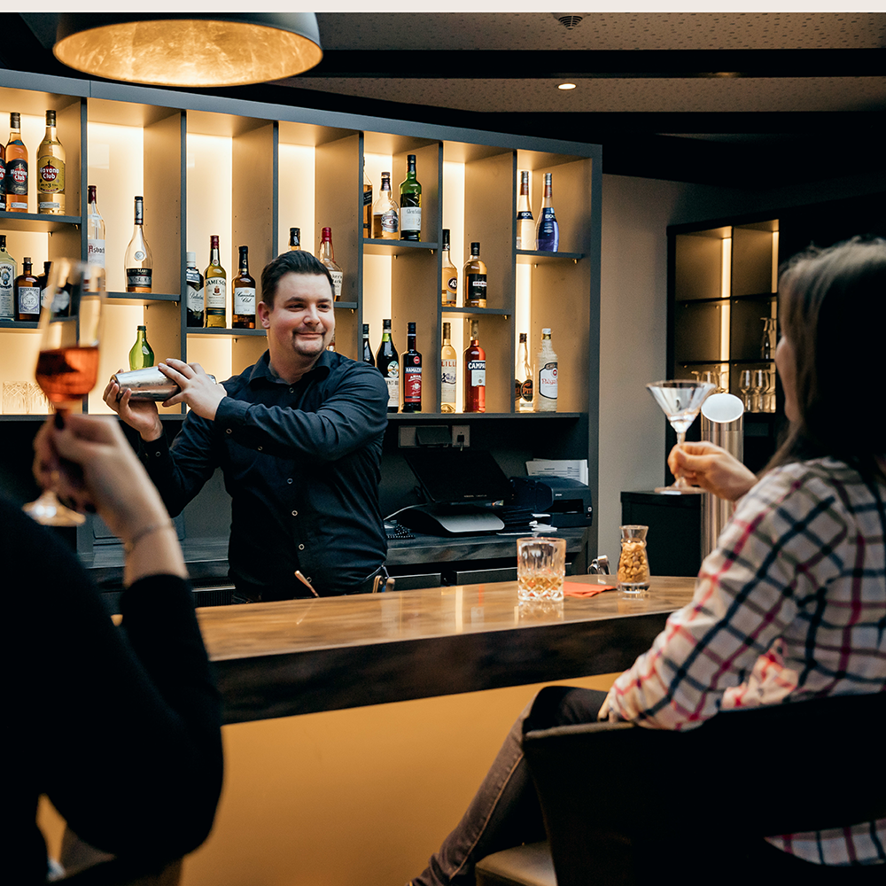 Barkeeper mixt Cocktail hinter einer Bar mit beleuchteten Regalen voller Spirituosen, Gäste halten Getränke in der Hand