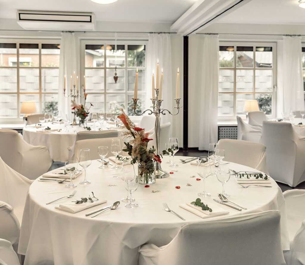 Rund gedeckter Tisch mit weißer Tischdecke, Kerzenleuchter und Blumendekoration in einem hellen, festlich eingerichteten Raum.