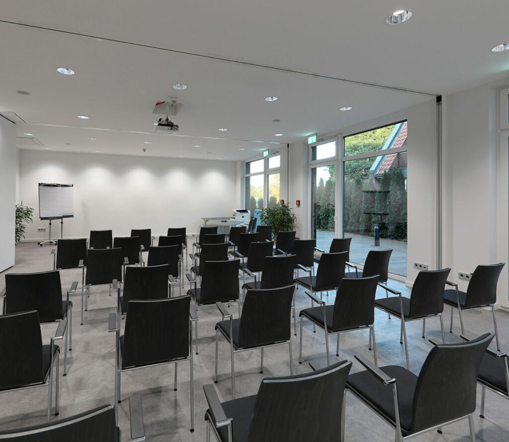Moderner Tagungsraum mit mehreren Reihen schwarzer Stühle, Flipchart und großen Fenstern mit Blick nach draußen.